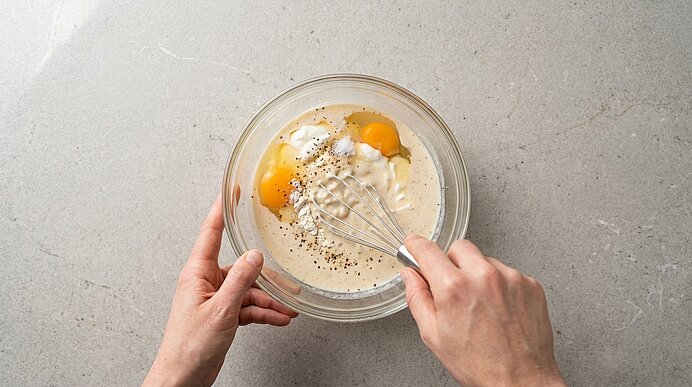 Zwei Hände verquirlen in einer Glasschüssel Eier mit Joghurt oder Sahne und Gewürzen mithilfe eines Schneebesens.