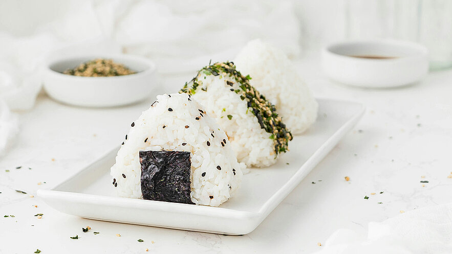 Drei Reisbällchen auf einem weißen Teller, jedes mit unterschiedlicher Garnitur. Eines hat ein schwarzes Nori-Blatt, das andere ist mit grünen Algen und schwarzen Sesamsamen bestreut. Im Hintergrund ist eine kleine Schüssel mit gewürztem Sesam und eine weitere Schüssel mit Sauce sichtbar.