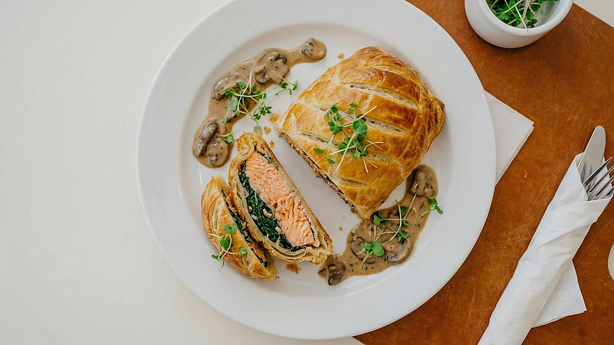 Ein Teller mit einem gefüllten Blätterteiggericht, das aufgeschnitten ist, um Lachs und Spinat zu zeigen. Um das Gericht herum sind kleine Tropfen einer cremigen Pilzsauce und einige frische Kräuter. Ein zusätzlicher Behälter mit Kräutern ist ebenfalls sichtbar.