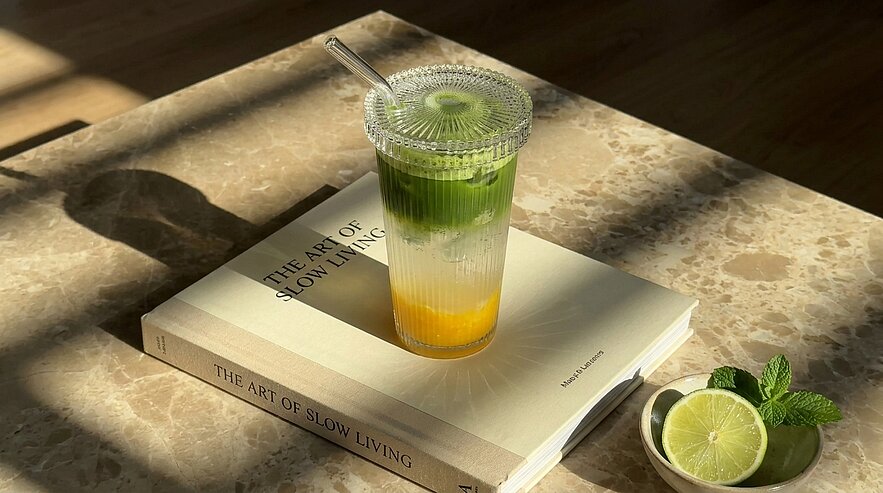 Klares Plastikglas mit Deckel und Strohhalm, geschichtetes Getränk in Grün, Weiß und Gelb, steht auf dem Buch „The Art of Slow Living“; daneben Limettenhälfte und Minzblätter im Sonnenlicht.