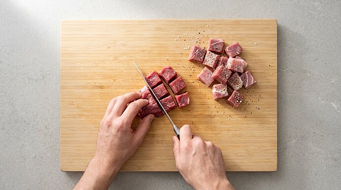 Hände schneiden rohes Rindfleisch in Würfel auf einem Holzbrett; daneben liegen bereits gewürfelte Fleischstücke, mit Pfeffer bestreut.