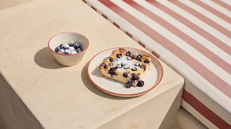 Stück Gebäck mit frischen Blaubeeren und Puderzucker auf einem Teller, daneben eine kleine Schale mit zusätzlichen Blaubeeren.