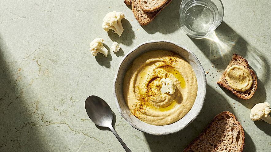 Eine Schüssel Blumenkohl-Hummus mit einem Spritzer Olivenöl ist zu sehen. Daneben befinden sich mehrere Scheiben Vollkornbrot und einige Blumenkohlröschen. Ein Esslöffel liegt neben der Schüssel, und ein Glas Wasser befindet sich im Hintergrund.