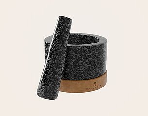 Ein grauer Mörser aus Stein mit einem passenden Stößel, auf einem hölzernen Untersetzer. Das Design ist schlicht und elegant, ideal zum Zerkleinern von Gewürzen oder Kräutern. Der Markenname "Waldwerk" ist auf dem Holz sichtbar.