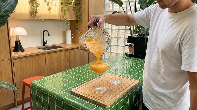 Person gießt orangen Smoothie aus Mixbehälter in zwei Cocktailgläser auf grün gekacheltem Küchentresen.