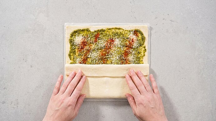 Zwei Hände drücken eine zweite Blätterteigschicht auf den mit Pesto, Parmesan und Paprikapulver belegten Teig.