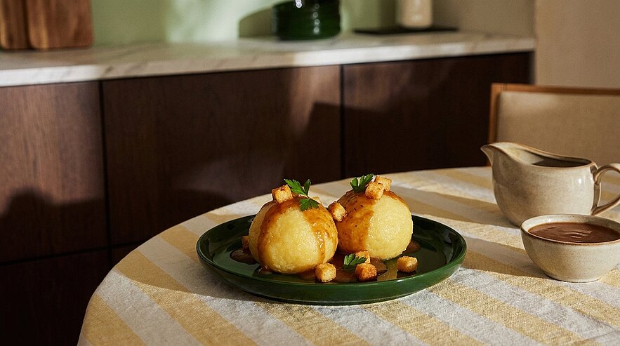 Zwei goldbraun frittierte Kugeln auf einem grünen Teller, garniert mit kleinen Croutons und frischen Kräuterblättern; daneben eine Schüssel und ein Soßenkännchen mit brauner Sauce.