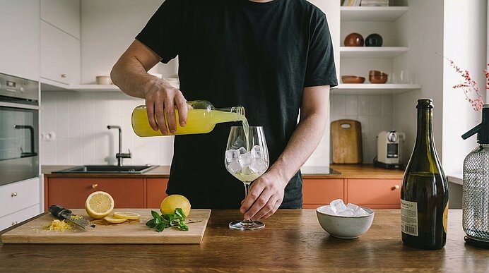 Person in schwarzem T‑Shirt gießt gelbe Zitronenlimonade aus einer Flasche in ein Weinglas mit Eis auf einer Küchenarbeitsplatte; Schneidebrett mit halbierten Zitronen, Messer und Minze.