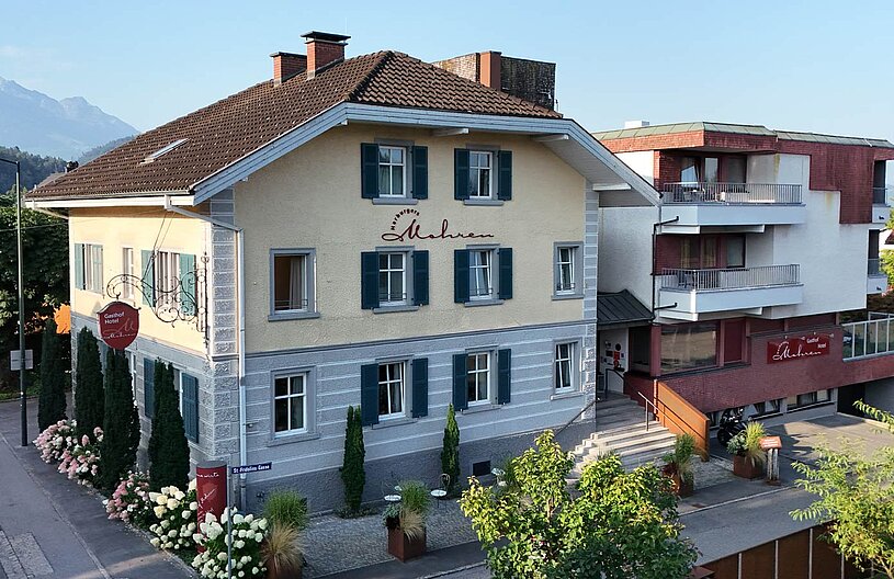 Eine bildschöne Gasthausansicht mit blauen Fensterläden und einem einladenden Eingangsbereich. Im Vordergrund stehen blühende Sträucher. Hinter dem Gasthaus sind majestätische Berge zu sehen. Der Himmel ist klar und die gesamte Szene vermittelt eine ruhige, einladende Atmosphäre.