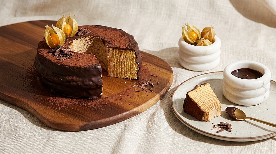 Runder, schokoladenüberzogener Baumkuchen mit sichtbaren feinen Schichten und einem herausgeschnittenen Stück auf einem Holzbrett; daneben eine Kuchenscheibe auf einem Teller, Schokoladensauce im kleinen Gefäß und dekorative Physalis.