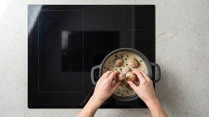 Obenaufnahme: Hände setzen mehrere Fleischbällchen in einen Topf mit cremiger Suppe auf einem schwarzen Kochfeld.