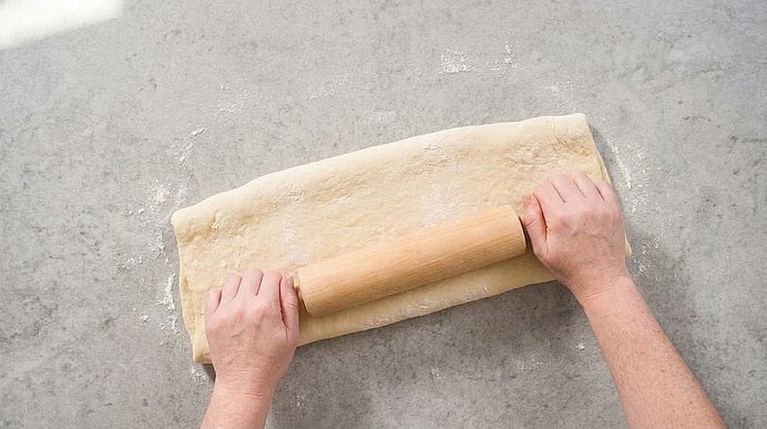 Zwei Hände rollen Teig mit einem hölzernen Nudelholz auf einer bemehlten hellgrauen Arbeitsfläche aus.