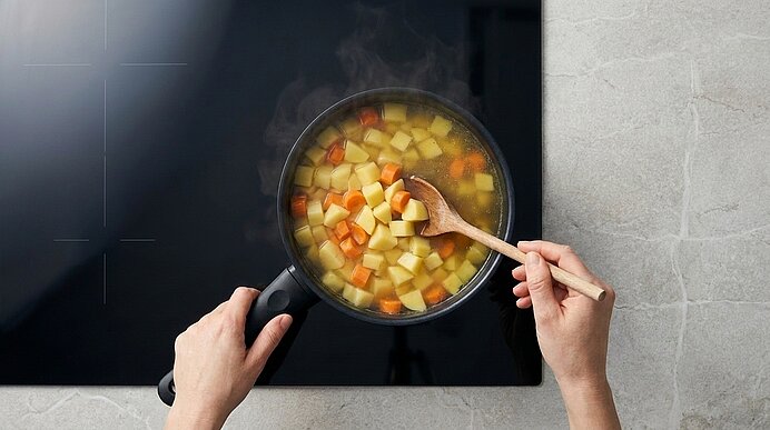Dampfender Topf mit Gemüsesuppe auf dem Herd, gewürfelte Kartoffeln und Karotten; eine Hand hält den Griff, die andere rührt mit einem Holzlöffel.