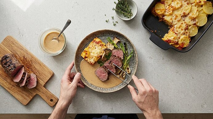 Hände schneiden rosa gebratenes Rindsteak auf einem Teller mit Bratensauce, dazu ein Stück Kartoffelgratin und grüne Bohnen; daneben Schneidebrett mit Braten und Auflaufform mit Gratin.