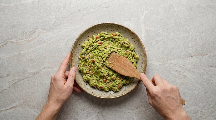 Zwei Hände streichen stückige Guacamole mit einem Holzspatel in einer Schale auf der hellgrauen Arbeitsplatte glatt.