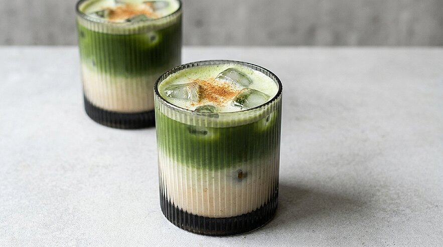 Zwei niedrige, geriffelte Gläser mit geschichtetem Eisgetränk: weiße Milchschicht unten, grüne Matcha-Schicht oben, Eiswürfel und eine Prise Zimt.
