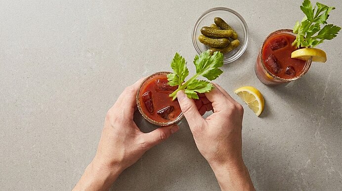 Hände halten ein Glas Bloody Mary mit Selleriestange und Zitronenscheibe; daneben ein zweites Glas und ein Schälchen Gewürzgurken.