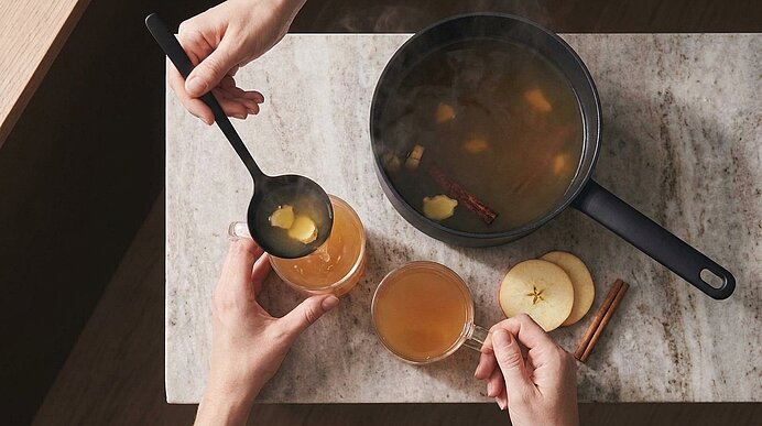 Zwei Hände schöpfen heißen Apfelpunsch aus einem Topf in ein Glas; im Topf sowie auf dem Tisch sichtbar: Apfelscheiben und Zimtstangen.