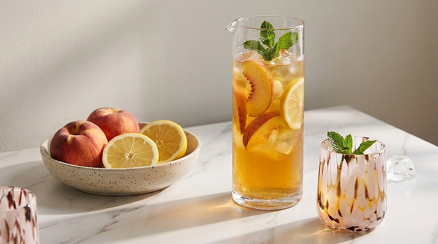 Krug mit Eistee, Eiswürfeln, Zitronen- und Pfirsichscheiben, garniert mit Minze; daneben ein Glas mit Eistee und Minze sowie eine Schale mit Pfirsichen und Zitronenscheiben auf hellem Tisch.
