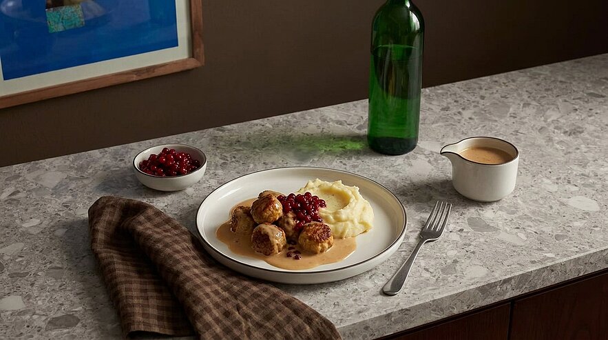 Teller mit schwedischen Fleischbällchen, Kartoffelpüree, brauner Soße und Preiselbeeren; daneben kleine Schale mit Preiselbeeren, Soßenkännchen und Gabel.
