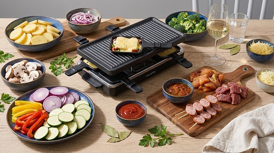 Eine Vielzahl von frischen Zutaten liegt auf einem Tisch, darunter geschnittene Kartoffeln, Brokkoli, Paprika, Zucchini, Zwiebeln, Pilze und Wurst. In der Mitte befindet sich ein Raclette-Grill mit einer Käseportion. Einige Schalen enthalten Dips. Zwei Gläser Weißwein stehen daneben.