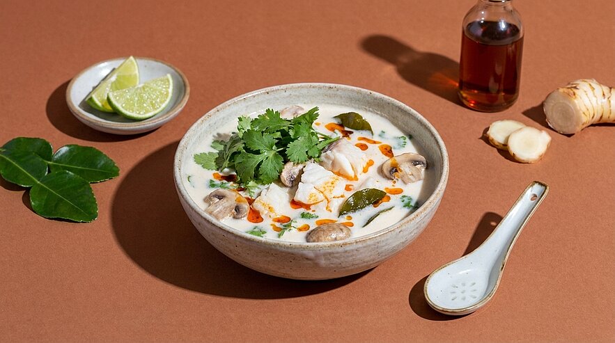 Schale mit cremiger Kokos-Hühnersuppe, garniert mit Champignons, frischem Koriander und Chiliflocken; Limettenstücke und ein Löffel neben der Schale.