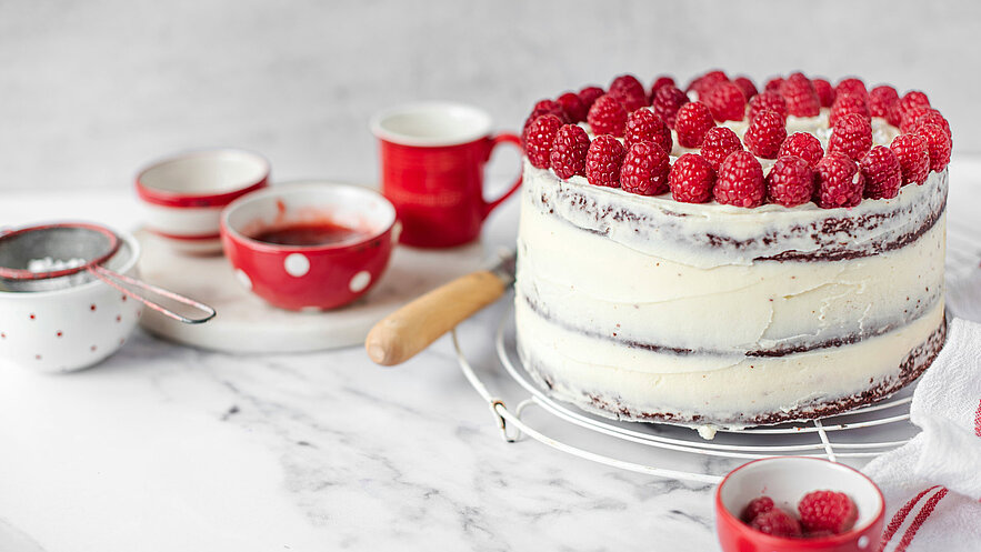 Eine zweistöckige Torte mit einer glatten weißen Buttercreme und frischen Himbeeren oben drauf. Rundherum sind einige rote Schalen und einen kleinen Sieb mit Zucker. Der Hintergrund ist unscharf und konzentriert sich auf die Torte im Vordergrund.