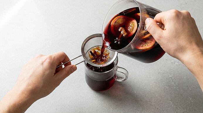 Zwei Hände gießen heißen Glühwein mit Orangenscheiben und Gewürzen aus einer Karaffe durch ein Sieb in eine Glastasse.