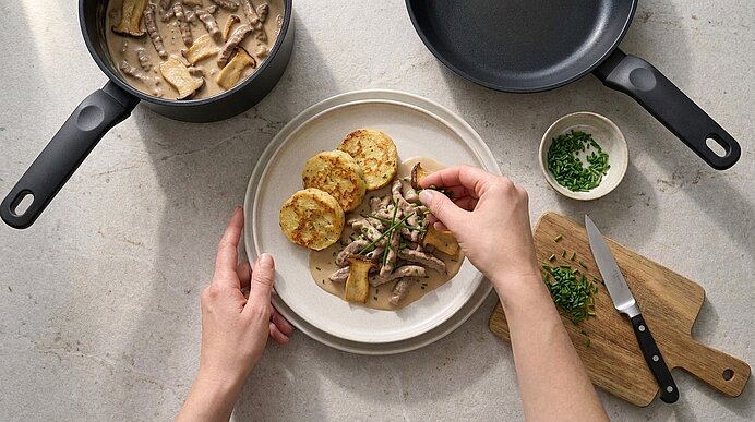 Hände garnieren einen Teller: cremiges Pilzragout mit geschnittenen Champignons neben drei Kartoffelplätzchen, mit frisch gehackten Kräutern bestreut.