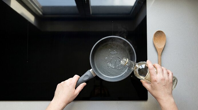 Zwei Hände gießen Öl aus einer Glasflasche in eine kleine Pfanne auf einem schwarzen Kochfeld; rechts liegt ein Holzlöffel.
