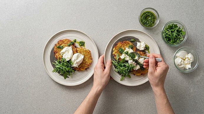 Hände beträufeln Kartoffelrösti, Burrata und Rucola auf einem Teller mit Kräuterpesto aus einem Löffel.