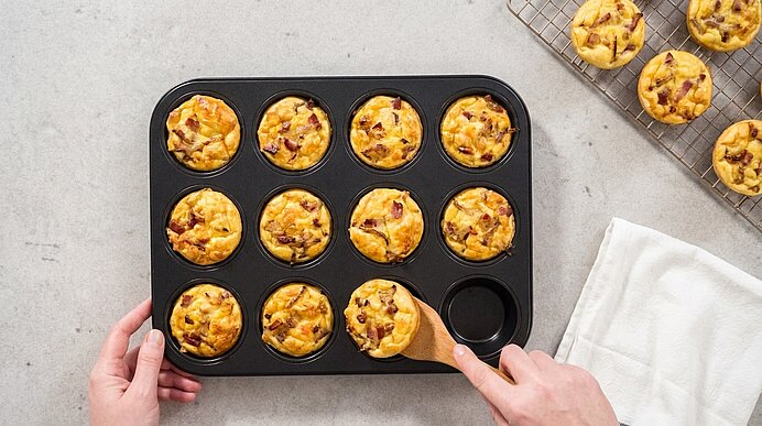 Zwei Hände heben eine fertig gebackene Mini-Quiche Lorraine mit einem Holzspatel aus einem schwarzen Muffinblech.