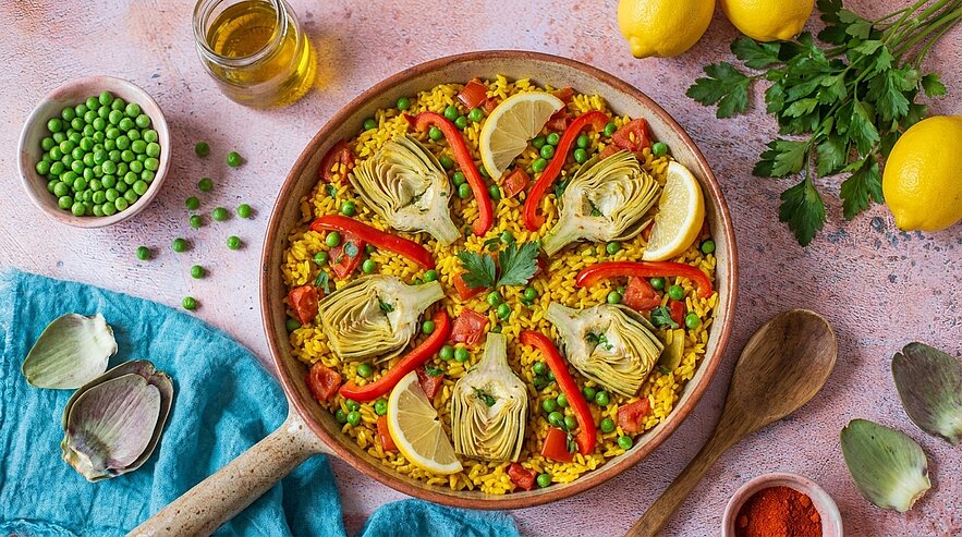 Eine große Pfanne mit farbenfrohem Reisgericht, dekoriert mit Artischocken, Paprika, Zitronenscheiben und Erbsen. Das Gericht ist umgeben von frischen Kräutern, Öl und einer sanften Tischdecke. Frische Zitronen liegen daneben, was dem Bild eine frische, einladende Atmosphäre verleiht.