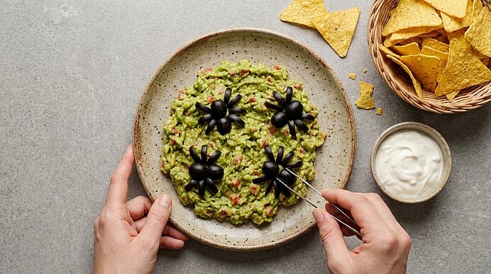 Zwei Hände legen Oliven-Spinnen mit einer Pinzette auf Guacamole, daneben stehen Sauerrahm und Tortilla-Chips.