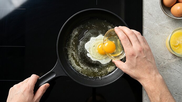 Hände halten eine Pfanne und gießen zwei aufgeschlagene Eier aus einer Glasschale in heißes Butterfett in der Pfanne.