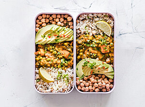 Zwei separate Fächer mit Reis und Kichererbsen. Im linken Fach befindet sich ein Gericht mit Gemüse und einer grünen Sauce, garniert mit Avocadoscheiben und Koriander. Im rechten Fach sind ebenfalls Reis und Kichererbsen, dazu eine Limettenscheibe.
