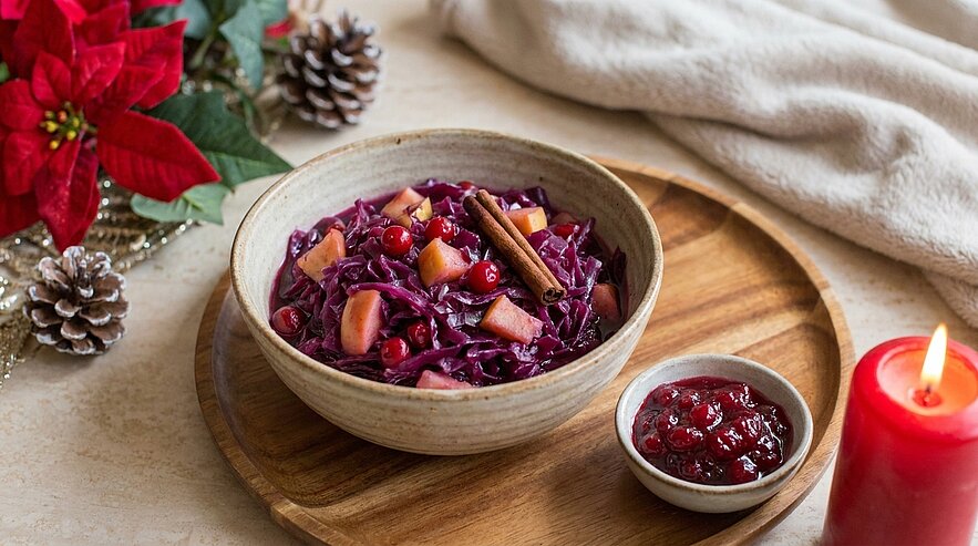 Schüssel geschmorter Rotkohl mit Apfelstücken, Preiselbeeren und einer Zimtstange, daneben eine kleine Schale Preiselbeersauce auf einem Holzbrett.