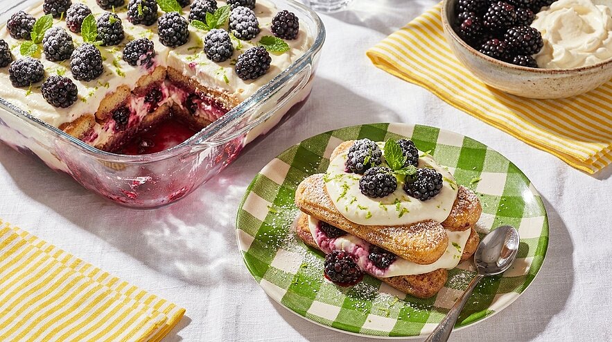 Glasschale mit geschichtetem Brombeer‑Dessert aus Biskuit, Beerenfüllung und Sahne; daneben ein Teller mit einem Stück, garniert mit frischen Brombeeren, Minze und Puderzucker.