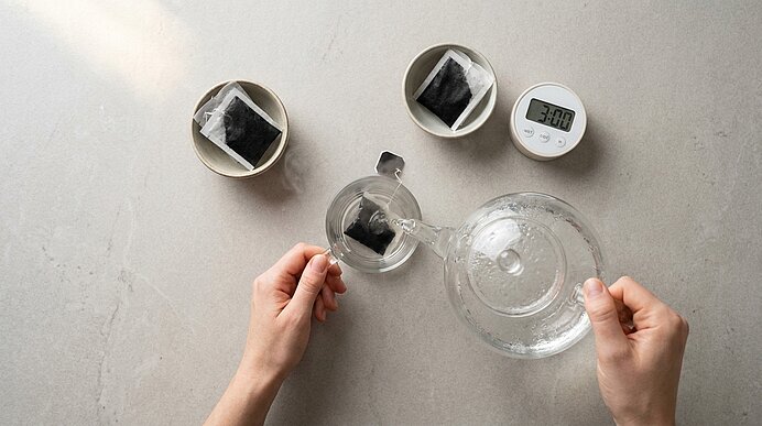 Zwei Hände gießen heißes Wasser aus einer Glaskanne in eine Tasse mit Teebeutel; daneben zwei weitere Tassen mit Teebeuteln und ein Timer mit Anzeige 3:00.