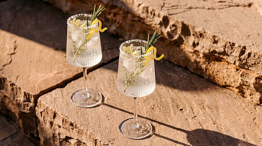 Zwei hohe Cocktailgläser mit prickelndem, eisgefülltem Getränk, gezuckertem Glasrand, je garniert mit Rosmarinzweig und Zitronenzeste, auf warmen Sandsteinstufen in der Sonne.