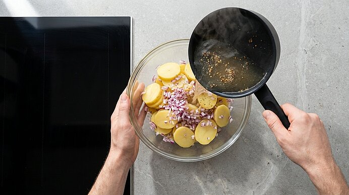 Zwei Hände gießen heißes Dressing aus einem kleinen Topf über eine Glasschüssel mit geschnittenen Kartoffelscheiben und fein gehackten roten Zwiebeln.