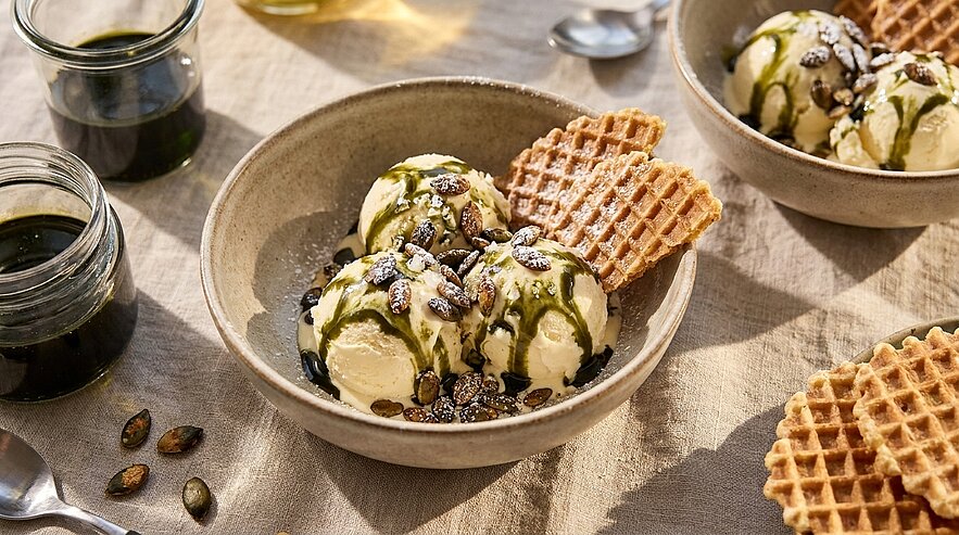 Schale mit drei Vanilleeiskugeln, beträufelt mit grünem Matcha- und dunklem Schokosirup, bestreut mit gerösteten Kürbiskernen; dazu eine knusprige Waffel.