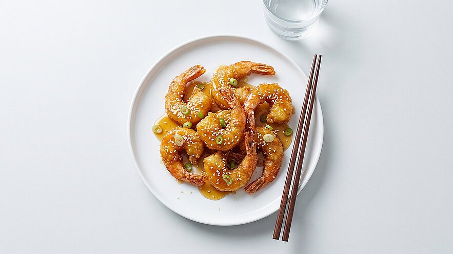 Ein Teller mit knusprigen, goldbraunen Shrimps in einer süß-sauren Sauce, garniert mit Sesam und frischen Frühlingszwiebeln. Zwei Essstäbchen liegen neben dem Teller.