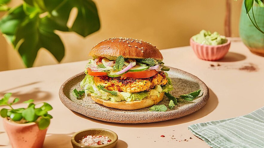 Ein saftiger Burger auf einem keramischen Teller, gefüllt mit gegrilltem Hähnchen, frischem Salat, Tomaten, Gurken, roten Zwiebeln und Minze. Der Brötchenoberteil ist mit Sesam bestreut. Im Hintergrund sind eine kleine Schale mit Gewürzen und eine Portion Avocadocreme sichtbar.
