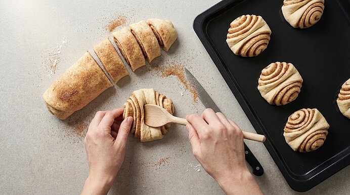 Hände formen Zimtschnecken aus einem geschnittenen Teigstrang, bestreut mit Zimt-Zucker; mehrere Schnecken liegen auf einem Backblech.