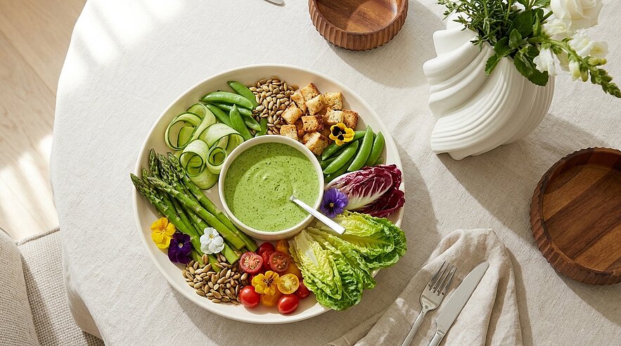 Schale mit grünem Dressing in der Mitte, umgeben von Spargel, Gurkenbändern, Zuckerschoten, Romanasalat, Radicchio, Kirschtomaten, Croutons, Samen und essbaren Blüten.