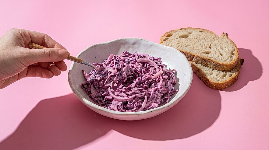 Hand rührt in einer Schale cremigen Rotkohlsalat; daneben liegen zwei Scheiben Bauernbrot auf pinkem Hintergrund.