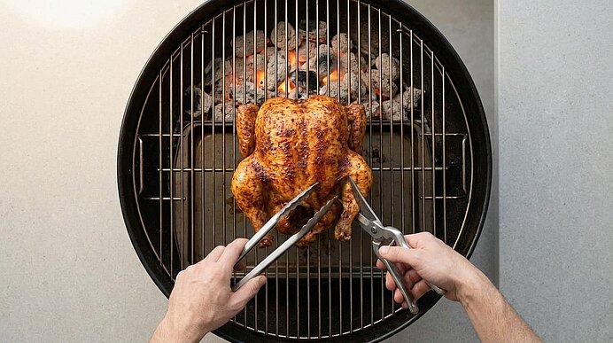 Zwei Hände schneiden ein knuspriges Brathähnchen auf dem Kugelgrill mit Geflügelschere und Grillzange.