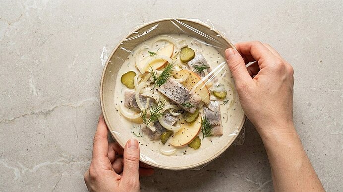 Hände ziehen Frischhaltefolie über eine Schale mit cremigem Heringssalat: Heringsstücke, Zwiebelringe, Apfelscheiben, Gewürzgurken und Dill.