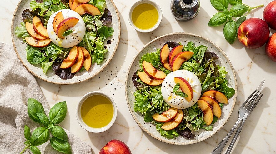 Zwei Teller Sommersalat: gemischte Blattsalate mit einer Burrata‑Kugel und Scheiben frischer Pfirsiche, garniert mit Basilikum, dazu kleine Schälchen Olivenöl.